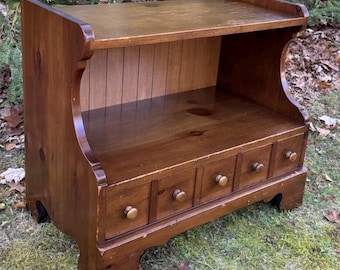 May include: A medium-brown wooden shelf unit featuring an open top storage area with a vertical paneled back and a base section containing four small drawers, each equipped with a single round wooden knob and curved side supports.