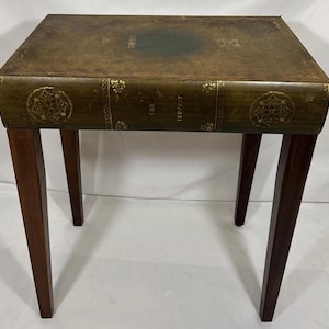 May include: A unique side table designed to resemble a large book. The table features a dark green leather-like top with gold lettering that reads "THE TEMPEST." It has four dark brown wooden legs.