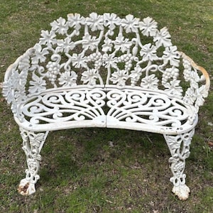 May include: A white cast metal bench featuring an intricate backrest decorated with grape clusters and vine leaves. The seat displays a symmetrical scrollwork pattern, and the four legs are adorned with detailed grape and leaf motifs.