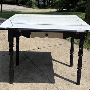 May include: A square table with a white top featuring a decorative black line border and a black apron. The table has four turned black legs and a single central drawer with a small black knob. The design includes a white drawer front panel.