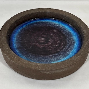 May include: A circular ceramic tray with a thick, matte dark brown rim and a smooth, glossy interior basin. The center features a deep black color that transitions into a vibrant, crackled electric blue ring near the outer edge.