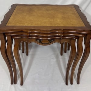May include: A set of three nested tables with dark brown wooden frames and curved legs. The largest table features a rectangular top with a tan, textured inlay. The middle table includes a small, ornate metal handle on the front apron.