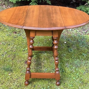 May include: A medium-brown wooden drop-leaf table with an oval top and two hinged side panels. The table features four turned legs connected by a rectangular base stretcher, providing a stable structure for the smooth, polished wooden surface.