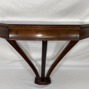 May include: A dark brown wooden wall-mounted console table featuring a smooth, flat top surface with beveled edges. The structure includes a central vertical support flanked by two angled, curved braces that meet at a small, octagonal base.