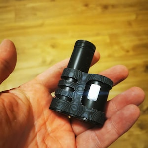 May include: Black and gray vaping device with a textured, patterned design. The device has a cylindrical top and a rectangular body with a small screen. The device is held in a hand.