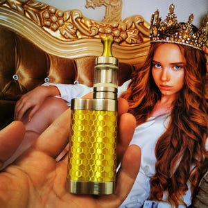 May include: A gold and silver vape pen with a honeycomb pattern. The pen is being held by a hand in front of a woman wearing a crown.