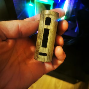 May include: A wooden vape mod with a rectangular body and three cut-out sections. The mod has a natural wood grain finish with dark brown streaks. The device is held in a hand, with a blurred background.