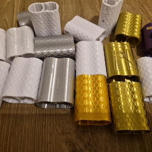 May include: A collection of cylindrical components in white, silver, gold, and purple. Some have textured surfaces with honeycomb or geometric patterns. These appear to be accessories or parts, possibly for a larger device or product.