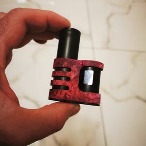 Puede incluir: Un mod de vapeo negro y rojo con un acabado texturizado similar a la veta de la madera. El dispositivo tiene un cuerpo rectangular con una parte superior negra y una pequeña pantalla. El mod se sostiene en una mano.