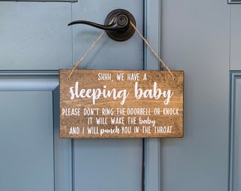 May include: A rectangular wooden sign with a dark brown stain and white text that reads: SHHH, WE HAVE A sleeping baby. PLEASE DON'T RING THE DOORBELL OR KNOCK. IT WILL WAKE THE baby AND I WILL punch YOU IN THE THROAT. The sign hangs by a twine cord.