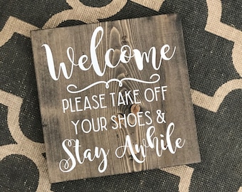 May include: A square wooden sign with the word "Welcome" in white script, followed by "PLEASE TAKE OFF YOUR SHOES & STAY AWHILE" in white block letters. The wood has a gray stain.