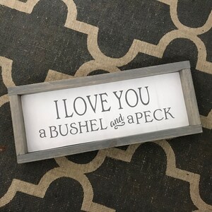 May include: A rectangular wooden sign with a gray frame and white background. The sign reads "I LOVE YOU a BUSHEL and a PECK" in black lettering. The sign is placed on a patterned gray and beige surface.