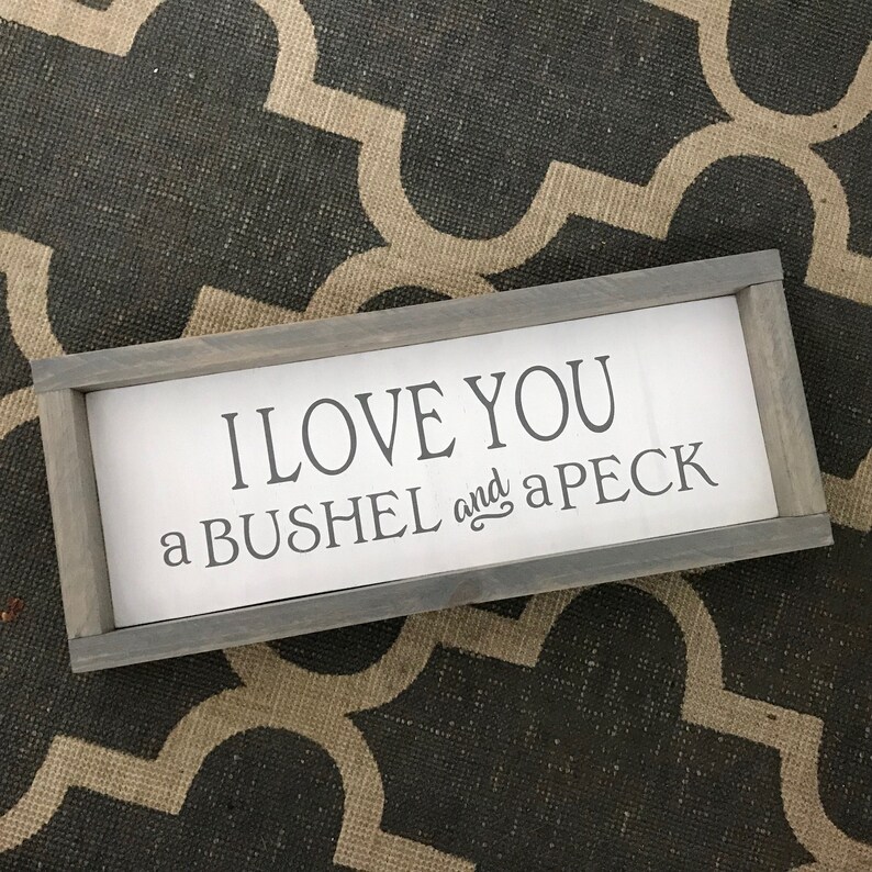 May include: A rectangular wooden sign with a gray frame and white background. The sign reads "I LOVE YOU a BUSHEL and a PECK" in black lettering. The sign is placed on a patterned gray and beige surface.