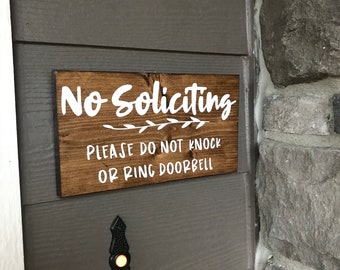 May include: A rectangular dark brown stained wooden sign with white text that reads No Soliciting, followed by a small leaf graphic, and the text PLEASE DO NOT KNOCK OR RING DOORBELL. The sign is mounted on a grey wall above a black doorbell button.