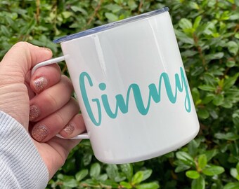 May include: White enamel mug with a teal-colored "Ginny" script. The mug has a white handle and a blue lid. The mug is held in front of a background of green foliage.