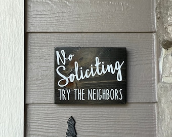 May include: A rectangular dark stained wooden sign with white lettering that reads No Soliciting in a script font and TRY THE NEIGHBORS in a clean sans-serif font. The sign is mounted on a horizontal siding wall above a black doorbell button.