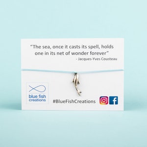 May include: A silver-colored dolphin charm bracelet. The charm hangs from a light blue cord attached to a white card with the text "The sea, once it casts its spell, holds one in its net of wonder forever" and "blue fish creations".