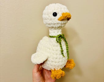 May include: A handmade white crochet goose with a yellow beak and feet. The goose has a green yarn bow around its neck and black eyes. The plush toy is soft and fluffy, perfect for a child's room or as a decorative item.