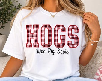 May include: White t-shirt with the word "HOGS" in large, red and black checkered letters. Below the word "HOGS" is the phrase "Woo Pig Sooie" in a black, cursive font. The shirt is a casual style.