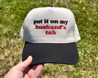 May include: A baseball cap with a black brim and a light gray crown. The text "put it on my husband's tab" is embroidered in red and black on the front of the cap.