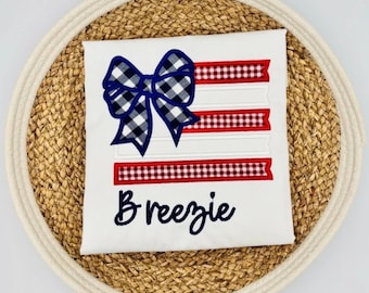 May include: White t-shirt with a patriotic design featuring a blue and white gingham bow and red and white striped ribbons. The name "Breezie" is embroidered in navy blue script. The shirt is displayed on a woven placemat.