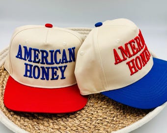 May include: Two baseball caps with the text "AMERICAN HONEY" embroidered on the front. One cap has a red brim and blue text, the other has a blue brim and red text. Both caps have a cream-colored crown and a button on top.
