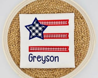May include: White t-shirt featuring a patriotic design with a blue and white checkered star and red and white striped accents. The name "Greyson" is embroidered in navy blue script below the design. The shirt is displayed on a woven placemat.
