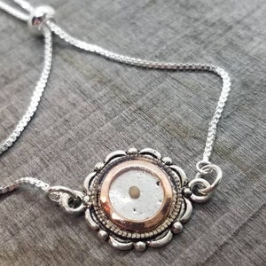 May include: A silver chain bracelet with a round pendant featuring a small, white seed encased in a clear resin. The pendant has a gold-toned rim and intricate detailing.