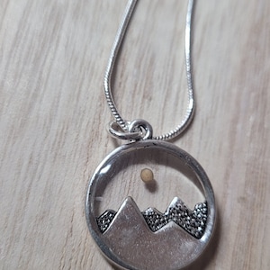 May include: A silver necklace with a round pendant featuring a mountain range silhouette and a small, round, beige stone in the center, representing the sun.