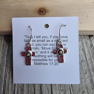 May include: Silver-toned cross earrings with a small, round, beige center. The earrings are displayed on a white card with a Bible verse from Matthew 17:20. The background is a wooden surface.