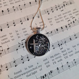 May include: A silver chain necklace with a round pendant featuring a black background with a silver glitter finish and a silver cross. The pendant is resting on a sheet of music with musical notes.