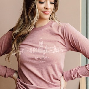 Puede incluir: Una mujer lleva una camiseta de manga larga de color rosa polvoriento con un gráfico blanco del horizonte de Londres. La camiseta está metida en unos vaqueros azules claros. El texto "London" es visible en el gráfico. La mujer mide 1,70 m y lleva una talla pequeña.