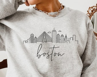 Op de afbeelding: Lichtgrijs sweatshirt met een zwarte lijntekening van de skyline van Boston, inclusief de Zakim Bridge, en het woord "Boston" in script eronder.