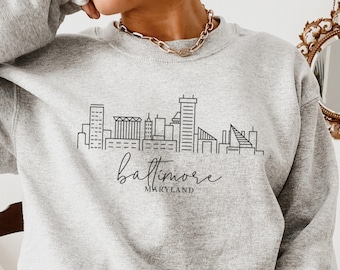 May include: A gray sweatshirt with a black outline of the Baltimore skyline and the text "Baltimore, Maryland".