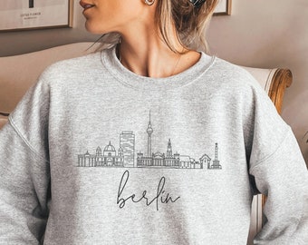 Puede incluir: Sudadera gris claro con un dibujo lineal negro del horizonte de Berlín. La palabra "berlin" está escrita en cursiva debajo. La sudadera es de estilo casual.