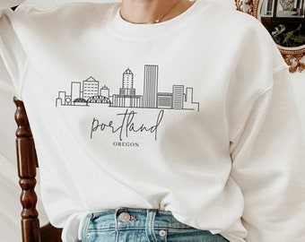 May include: White sweatshirt with a black outline of the Portland skyline and the text "portland OREGON". A person is wearing the sweatshirt and blue jeans.