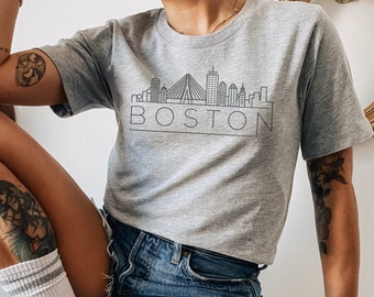 May include: A gray t-shirt with a black outline of the Boston skyline and the word "BOSTON" printed on the front. The shirt is being worn by a person with a tattoo on their leg.