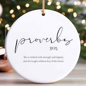 May include: A white, circular Christmas ornament featuring the word "proverbs" in black script. The text "31:25" and a quote about strength and dignity are below. A gold string is attached for hanging. Christmas tree and baubles are in the background.