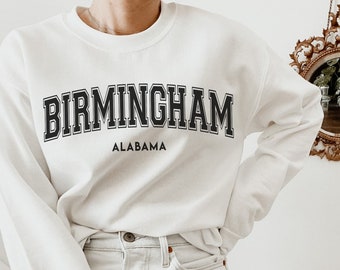 以下が含まれることがあります： 「BIRMINGHAM」の文字が黒いブロック体で「ALABAMA」の上にプリントされた白いスウェットシャツ。明るい色のデニムショーツと組み合わせています。背景には、金色のフレームの装飾的な鏡が掛けられています。