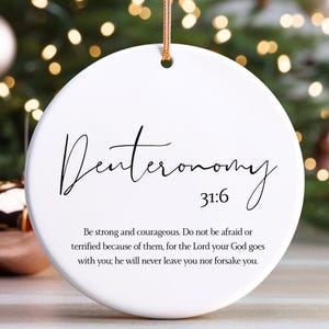 May include: A white, round Christmas ornament with the word "Deuteronomy" in elegant script. The ornament also features the verse "31:6" and a longer inspirational quote. A gold string is attached for hanging.