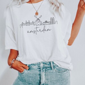 May include: White t-shirt featuring a black line drawing of the Amsterdam skyline, including a windmill, buildings, and a bridge. The text "amsterdam" is written below the skyline.