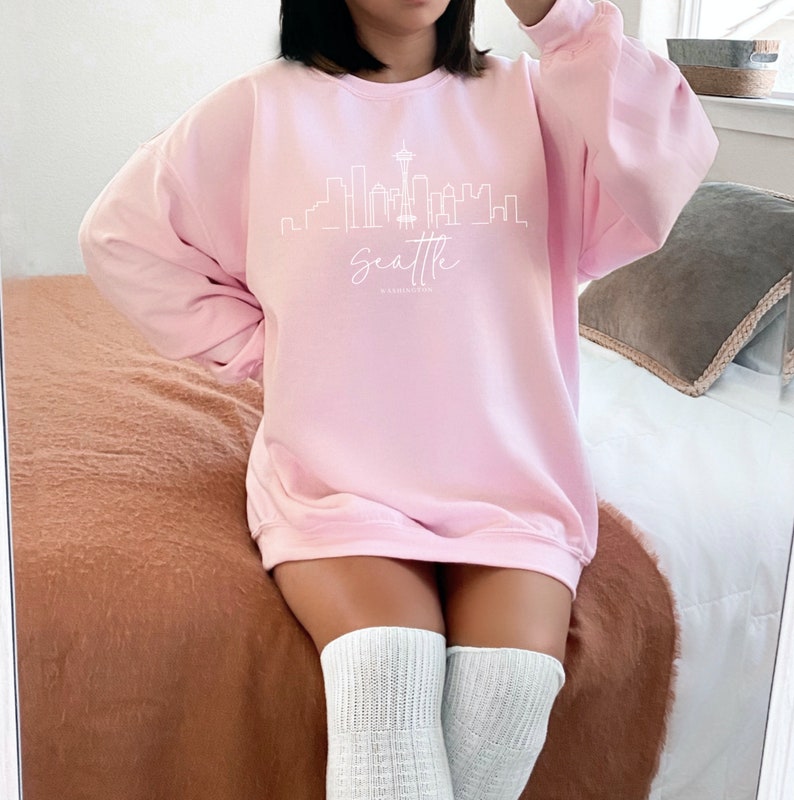 Pode incluir: Uma camisa de moletom rosa com um contorno branco do horizonte de Seattle e o texto "Seattle, Washington" impresso na frente.