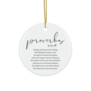 May include: A white, round ceramic ornament with the word "proverbs" in a cursive font. Below the word is the text "31:25-28" and a longer passage of text. A gold string is attached for hanging.
