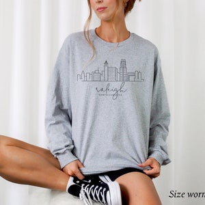 May include: Gray sweatshirt with a black outline of the Raleigh, North Carolina skyline and the text "Raleigh, North Carolina". The model is wearing a size large.