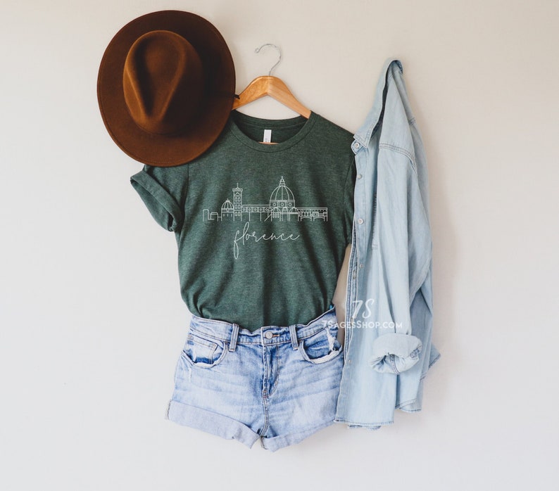May include: A green t-shirt with a white outline of the Florence skyline and the word "Florence" written in cursive. The shirt is hanging on a wooden hanger with a brown hat and a light blue denim jacket.