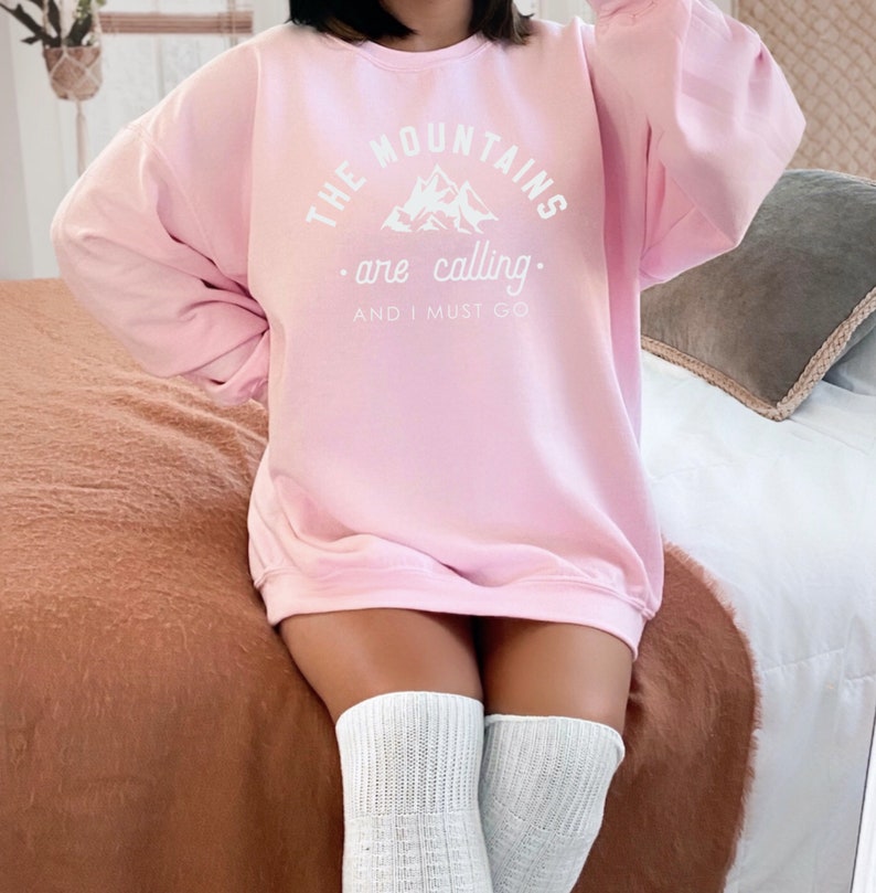 May include: A pink sweatshirt with the text "THE MOUNTAINS are calling. AND I MUST GO" in white. The text is surrounded by a mountain graphic.