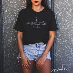 K&ouml;nnte beinhalten: Schwarzes T-Shirt mit einer minimalistischen Strichzeichnung der Londoner Skyline, einschlie&szlig;lich Big Ben und Tower Bridge. Das Wort "London" steht in Schreibschrift darunter, darunter "England". Das T-Shirt ist in Gr&ouml;&szlig;e Large.