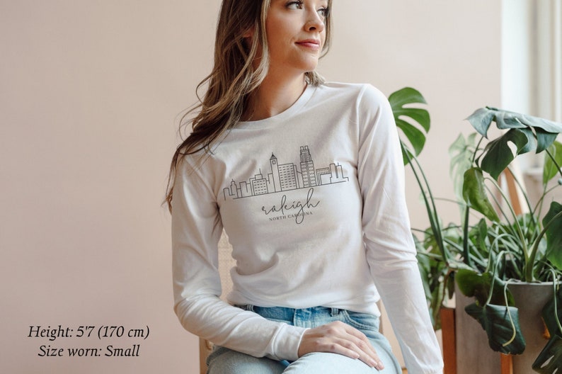 May include: White long-sleeve t-shirt with a black outline of the Raleigh, North Carolina skyline and the text "raleigh north carolina".