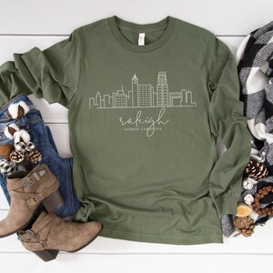 May include: A green long-sleeved t-shirt with a white outline of the Raleigh, North Carolina skyline and the text "raleigh NORTH CAROLINA".