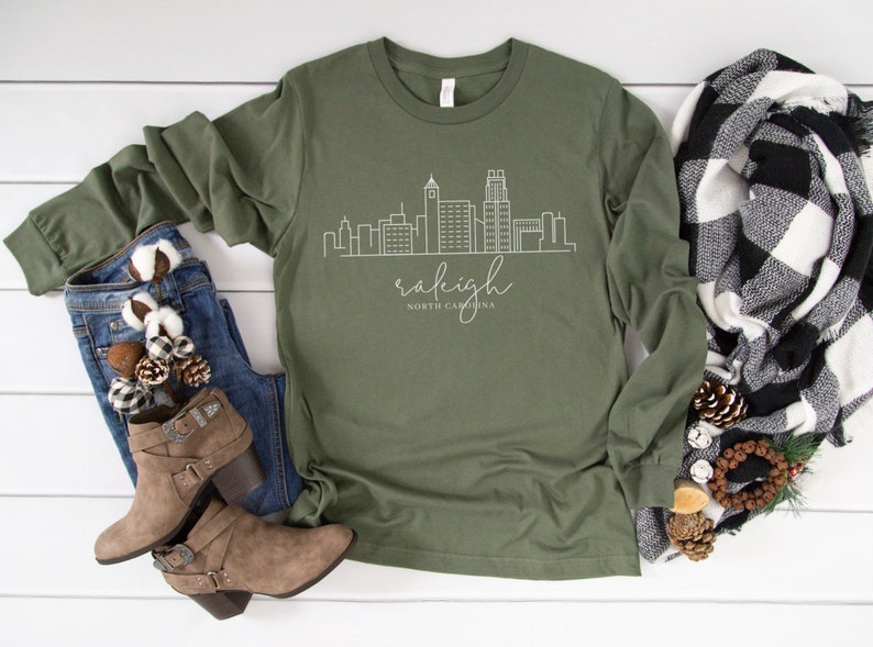 May include: A green long-sleeved t-shirt with a white outline of the Raleigh, North Carolina skyline and the text "raleigh NORTH CAROLINA".